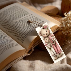 May include: A bookmark featuring two teddy bears, one hugging the other, both holding bouquets of flowers. The bears wear pink and white outfits and are adorned with floral accents. A brown string is attached to the top of the bookmark.