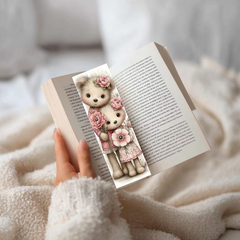 May include: A decorative bookmark featuring two teddy bears with floral accents. The bears are dressed in pink outfits and holding flowers. The bookmark is inserted into an open book, with the page visible.