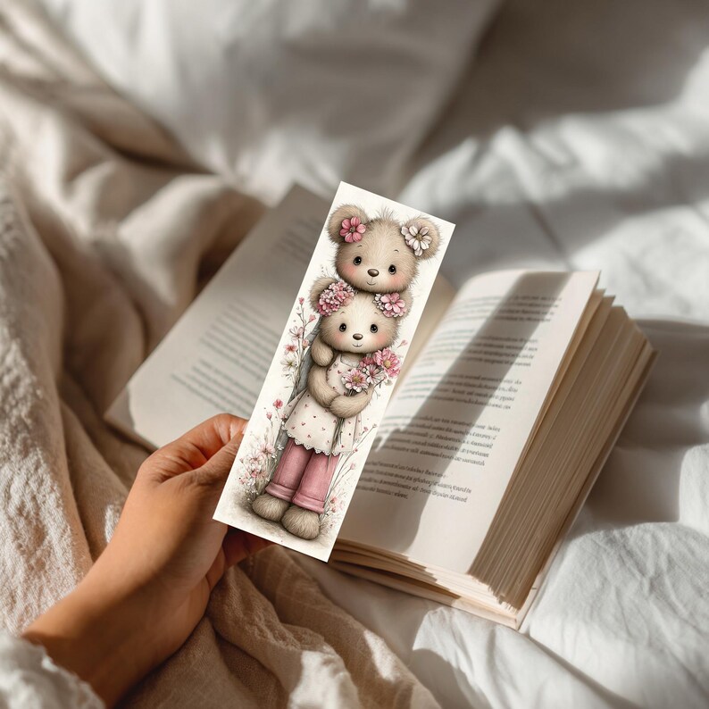 May include: A whimsical bookmark featuring two teddy bears adorned with floral accents. The top bear has a flower crown, while the bottom bear holds a bouquet. The bookmark is held over an open book, suggesting a reading theme.