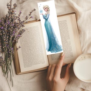 Peut inclure: Un marque-page en papier rectangulaire pr&eacute;sentant une illustration imprim&eacute;e d'un personnage blond avec une coiffure tress&eacute;e, portant une longue robe bleue scintillante et tenant un flocon de neige bleu sur un fond blanc avec des flocons.