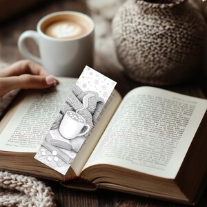 May include: A black and white bookmark featuring a cozy illustration of a mug of hot cocoa, a knitted scarf, and books. The bookmark is placed in an open book, with a cup of coffee in the background. The theme is reading and comfort.