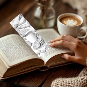 May include: A black and white bookmark featuring a detailed illustration of a mug of coffee, cookies, and a rolling pin. The bookmark is placed in an open book, next to a cup of coffee with latte art. The scene evokes a cozy, reading-themed atmosphere.