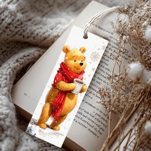 Peut inclure: Un marque-page rectangulaire en papier illustr&eacute; d'un ours jaune portant une &eacute;charpe rouge vif et tenant une tasse blanche. L'illustration comporte de petits flocons de neige blancs sur fond blanc, avec une fine ficelle blanche attach&eacute;e en haut.