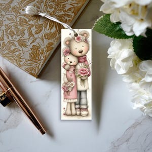 May include: A decorative bookmark featuring two cartoon teddy bears. The bears are adorned with floral accents and hold bouquets of pink flowers. One bear wears a pink dress, and the other wears a scarf. The bookmark has a white tassel.