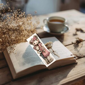 May include: A bookmark featuring a watercolor illustration of two teddy bears, one larger and one smaller, both adorned with floral crowns and holding flowers. The bears are dressed in pink and gray outfits. The bookmark is placed in an open book.