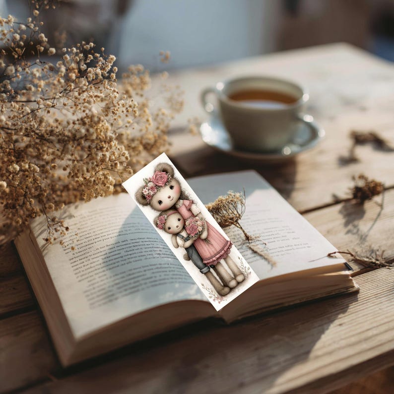 May include: A bookmark featuring a watercolor illustration of two teddy bears, one larger and one smaller, both adorned with floral crowns and holding flowers. The bears are dressed in pink and gray outfits. The bookmark is placed in an open book.