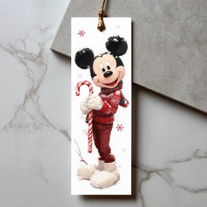 Peut inclure: Un marque-page rectangulaire illustr&eacute; avec Mickey Mouse portant un pull rouge &agrave; motifs, une &eacute;charpe rouge et verte, et des bottes blanches. Mickey tient une canne en sucre rouge et blanche, entour&eacute; de petits flocons rouges sur fond blanc.