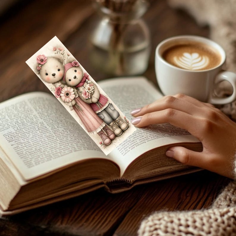 May include: A decorative bookmark featuring two cartoon teddy bears, one in a pink dress and the other in a red scarf and jacket, holding flowers. The bookmark is placed in an open book, with a cup of coffee in the background.