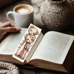 May include: A whimsical bookmark featuring two teddy bears, one with a floral crown and the other with a bow. The bears are holding flowers and wearing pink outfits. The bookmark is placed in an open book, with a cup of coffee in the background.