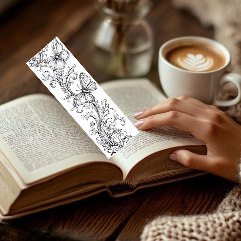 Op de afbeelding: Een rechthoekige boekenlegger met een zwart-witte lijntekening van twee vlinders omringd door ingewikkelde, krullende bloemen- en rankpatronen. De boekenlegger is verticaal tussen de pagina's van een open boek geplaatst.