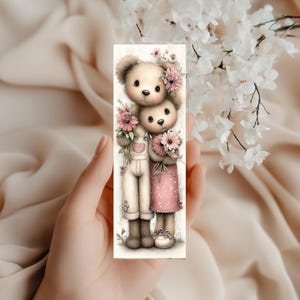 May include: A rectangular bookmark featuring two teddy bears with floral accents. One bear wears overalls with a heart, the other a pink dress. Both hold bouquets of pink flowers. The background is a soft beige.
