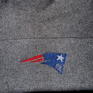 May include: A grey knitted beanie hat with an embroidered team logo in red, white, and blue. The logo is a stylized flag design with a white star. The hat is made of a soft, textured material.