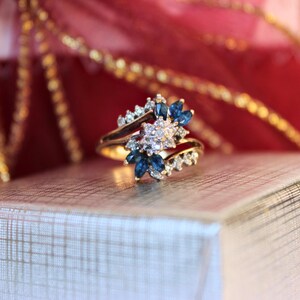 May include: A gold ring with a floral design, featuring blue and clear gemstones. The ring is set against a backdrop of red and gold, suggesting a gift or special occasion. The gemstones are arranged to resemble petals.