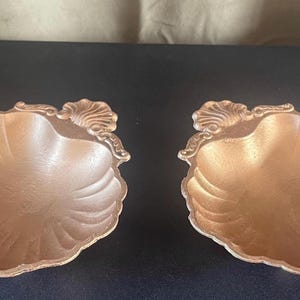 May include: Two decorative, seashell-shaped dishes with a metallic bronze finish. The dishes have a scalloped edge and a detailed, fan-like pattern radiating from the center. The design includes a decorative shell accent on one side.