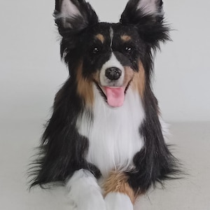 May include: A plush dog with black, white, and brown fur, posed with its tongue out. The dog has large, upright ears and is lying down with its front paws extended. The dog's fur is long and fluffy, and its eyes are open.