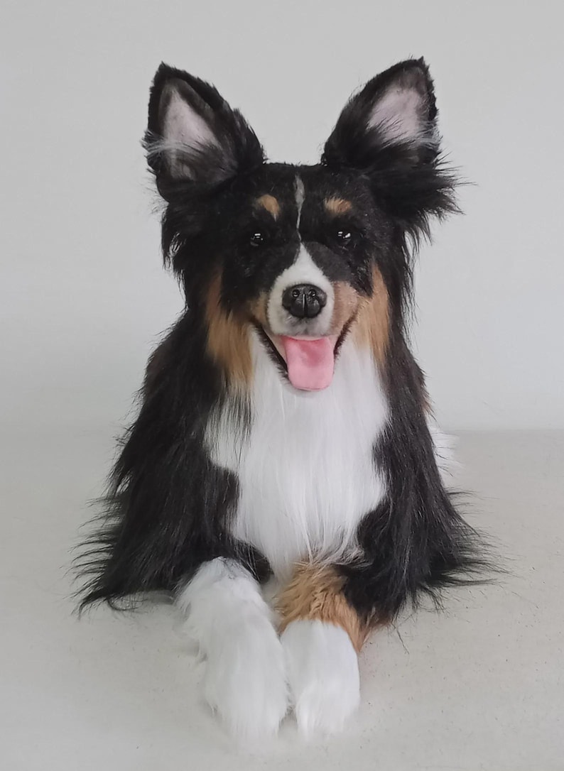 May include: A plush dog with black, white, and brown fur, posed with its tongue out. The dog has large, upright ears and is lying down with its front paws extended. The dog's fur is long and fluffy, and its eyes are open.