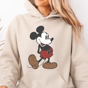May include: Sand-colored hoodie featuring a large graphic of Mickey Mouse. The cartoon character is depicted in his classic outfit: red shorts, brown shoes, and black gloves. The hoodie has a front pocket and a drawstring hood.