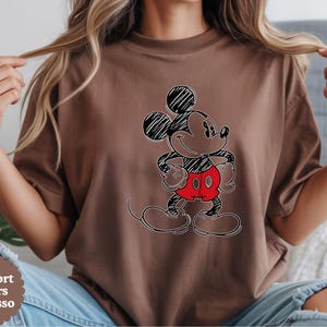 May include: Espresso-colored t-shirt featuring a sketched Mickey Mouse design. The cartoon character is depicted with black outlines, red shorts, and a smiling expression. The shirt's color is indicated by the text "Comfort Colors Espresso" in a circular design.