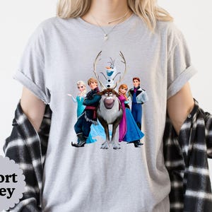 May include: A light grey short-sleeve t-shirt featuring a graphic print of five animated characters including a snowman, a reindeer, and three people in colourful outfits. The shirt is layered under a black and white plaid button-down shirt.