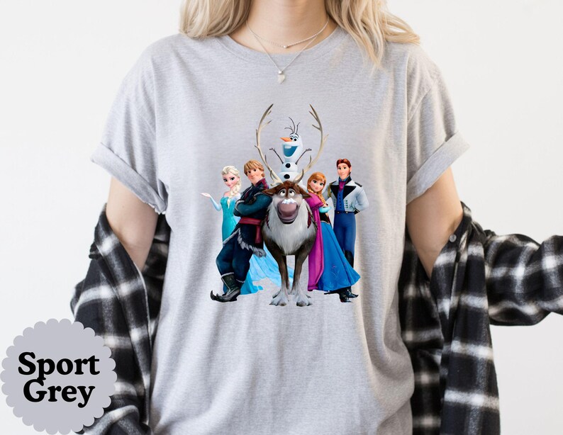 May include: A light grey short-sleeve t-shirt featuring a graphic print of five animated characters including a snowman, a reindeer, and three people in colourful outfits. The shirt is layered under a black and white plaid button-down shirt.