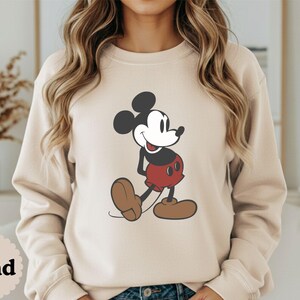May include: Sand-colored sweatshirt featuring a classic Mickey Mouse graphic. The cartoon character is depicted in black, red, and brown, with a smiling expression. The word "Sand" is printed at the bottom left of the image.