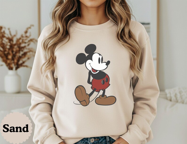 May include: Sand-colored sweatshirt featuring a classic Mickey Mouse graphic. The cartoon character is depicted in black, red, and brown, with a smiling expression. The word "Sand" is printed at the bottom left of the image.