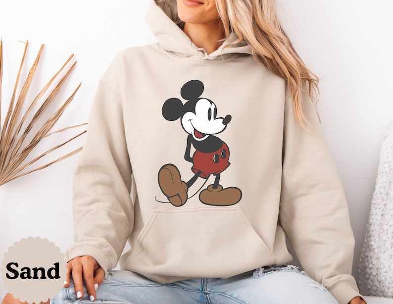 May include: Sand-colored hoodie featuring a large graphic of Mickey Mouse. The cartoon character is depicted in his classic outfit: red shorts, brown shoes, and black gloves. The hoodie has a front pocket and a drawstring hood.