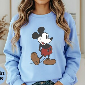 May include: Light blue sweatshirt featuring a classic Mickey Mouse graphic. The cartoon character is depicted in his iconic pose, wearing red shorts, brown shoes, and a black and white body. The text "Light Blue" is in a blue circle at the bottom.