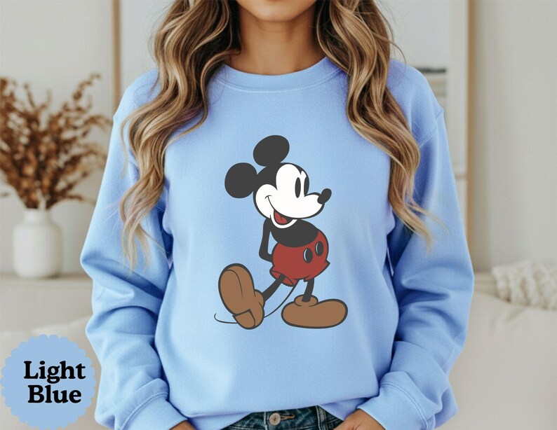 May include: Light blue sweatshirt featuring a classic Mickey Mouse graphic. The cartoon character is depicted in his iconic pose, wearing red shorts, brown shoes, and a black and white body. The text "Light Blue" is in a blue circle at the bottom.