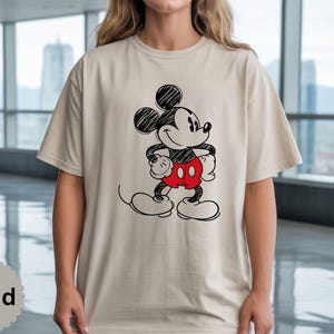 May include: Sand-colored t-shirt featuring a black and red sketch of Mickey Mouse. The cartoon character is depicted with his signature large ears, red shorts, and white gloves and shoes. The shirt is a casual, unisex style.