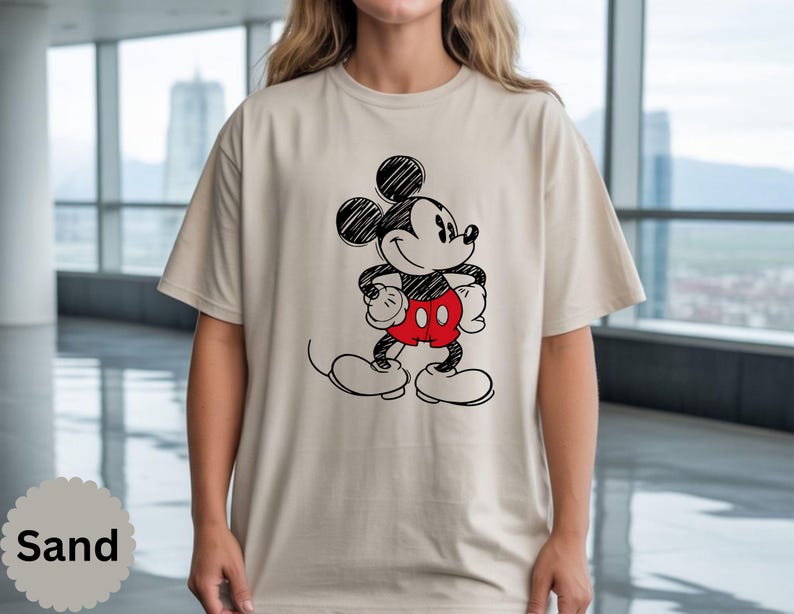 May include: Sand-colored t-shirt featuring a black and red sketch of Mickey Mouse. The cartoon character is depicted with his signature large ears, red shorts, and white gloves and shoes. The shirt is a casual, unisex style.