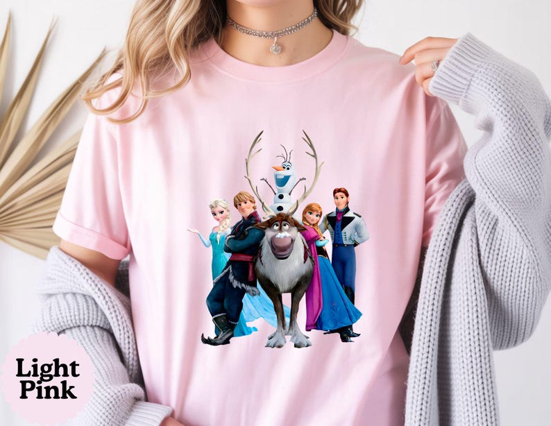 May include: A light pink short-sleeved t-shirt featuring a central graphic print of the animated characters Elsa, Kristoff, Olaf, Sven, Anna, and Hans. The crew neck shirt is made of a solid-coloured fabric and displays the characters in full colour.