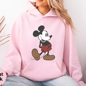 May include: Light pink hoodie featuring a classic Mickey Mouse graphic. The cartoon character is depicted in black, red, and brown. The hoodie has a kangaroo pocket and a drawstring hood. The text "Light Pink" is visible in a pink circle.