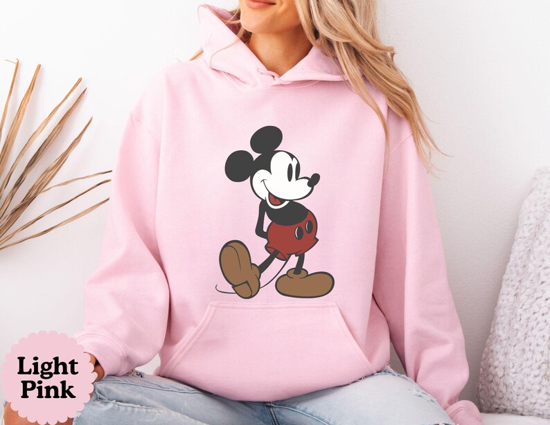 May include: Light pink hoodie featuring a classic Mickey Mouse graphic. The cartoon character is depicted in black, red, and brown. The hoodie has a kangaroo pocket and a drawstring hood. The text "Light Pink" is visible in a pink circle.