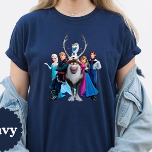 May include: A navy blue short-sleeve t-shirt featuring a graphic print of the characters Elsa, Kristoff, Olaf, Sven, Anna, and Hans from the movie Frozen. The shirt has a crew neckline and is worn with a light blue denim jacket over the shoulders.