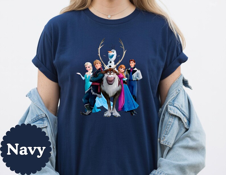 May include: A navy blue short-sleeve t-shirt featuring a graphic print of the characters Elsa, Kristoff, Olaf, Sven, Anna, and Hans from the movie Frozen. The shirt has a crew neckline and is worn with a light blue denim jacket over the shoulders.