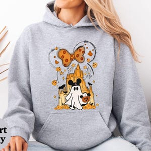 May include: A sport grey hooded sweatshirt featuring a graphic print of a ghost wearing mouse ears, standing in front of a castle. The design includes an orange polka-dot bow, small pumpkins, bats, and stars. The text Sport Grey is visible in a circle.