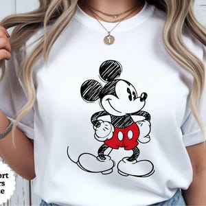 May include: White t-shirt featuring a black and white sketch of Mickey Mouse with red shorts. The text "Comfort Colors White" is in a white oval on the bottom left. The shirt is a classic crew neck.
