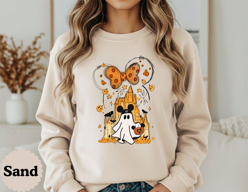 May include: A sand-colored crewneck sweatshirt featuring a graphic print of a ghost wearing mouse ears in front of a castle. The design includes an orange polka-dot bow, small pumpkins, stars, and bats in shades of orange, black, and yellow.