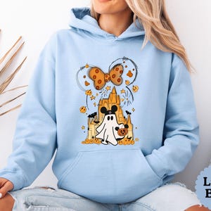 May include: A light blue hooded sweatshirt featuring a graphic print of a ghost wearing mouse ears, standing in front of a castle. The design includes a large orange bow, small pumpkins, stars, and bats. The hoodie has long sleeves and a front pocket.