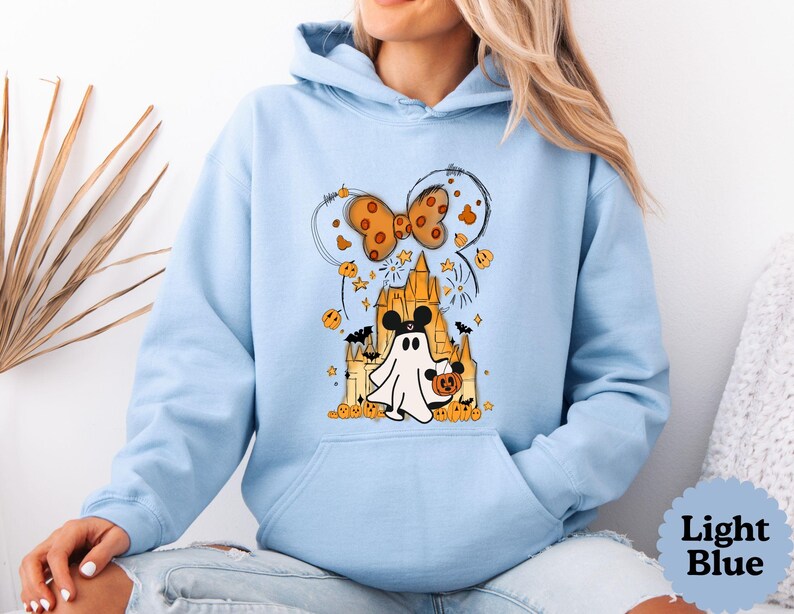 May include: A light blue hooded sweatshirt featuring a graphic print of a ghost wearing mouse ears, standing in front of a castle. The design includes a large orange bow, small pumpkins, stars, and bats. The hoodie has long sleeves and a front pocket.
