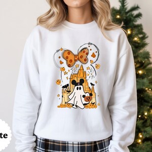 May include: A white long-sleeve sweatshirt featuring a graphic print of a ghost wearing mouse ears, holding a pumpkin bucket in front of a castle. Above the castle is a large orange polka-dot bow, surrounded by small pumpkins, stars, and bats.