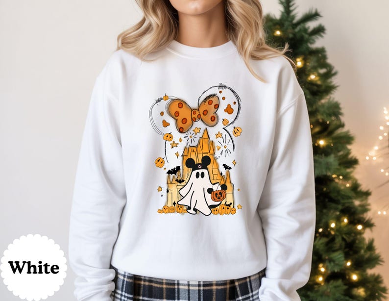 May include: A white long-sleeve sweatshirt featuring a graphic print of a ghost wearing mouse ears, holding a pumpkin bucket in front of a castle. Above the castle is a large orange polka-dot bow, surrounded by small pumpkins, stars, and bats.