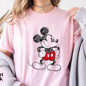 May include: Light pink t-shirt featuring a black and white sketch of Mickey Mouse with red shorts. The text "Light Pink" is in the bottom left corner. The shirt is a crew neck and has short sleeves.