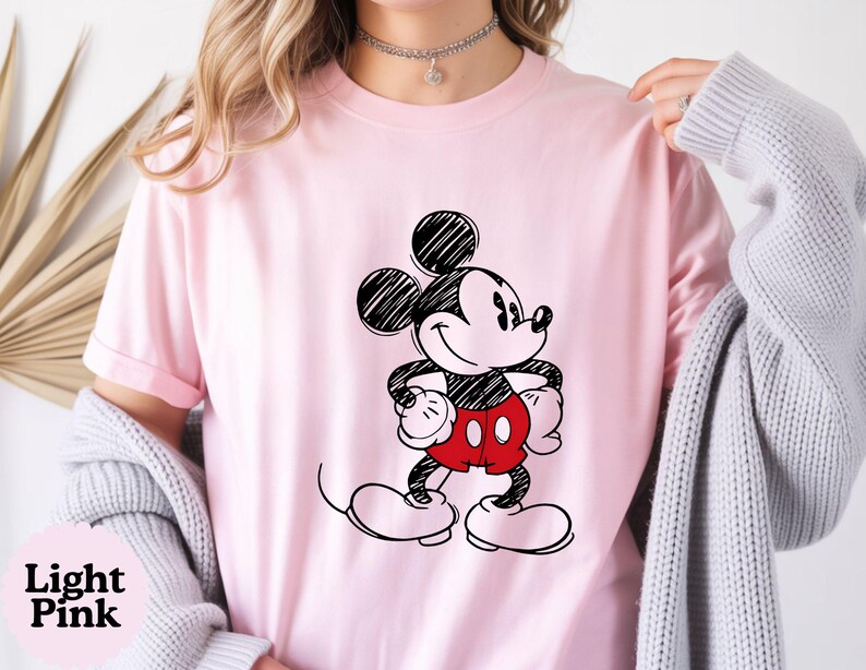 May include: Light pink t-shirt featuring a black and white sketch of Mickey Mouse with red shorts. The text "Light Pink" is in the bottom left corner. The shirt is a crew neck and has short sleeves.