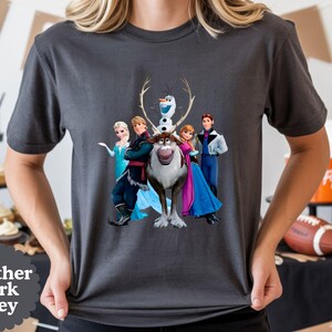 May include: A heather dark grey short-sleeved t-shirt featuring a graphic print of the characters Elsa, Kristoff, Olaf, Sven, Anna, and Hans from the movie Frozen. The design is centred on the chest of the shirt, displaying the characters together.