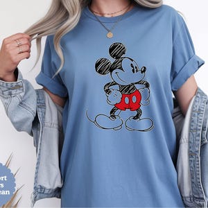 May include: A blue Comfort Colors t-shirt featuring a black and red Mickey Mouse graphic. The cartoon character is smiling with his hands on his hips. The shirt has rolled-up sleeves and a relaxed fit.
