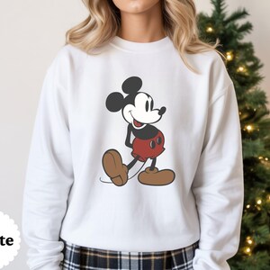 May include: White sweatshirt featuring a classic Mickey Mouse graphic. The cartoon character is depicted in his iconic pose, wearing red shorts, brown shoes, and black gloves. The sweatshirt has a crew neck and long sleeves. The word "White" is printed on the bottom left.