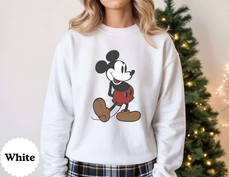 May include: White sweatshirt featuring a classic Mickey Mouse graphic. The cartoon character is depicted in his iconic pose, wearing red shorts, brown shoes, and black gloves. The sweatshirt has a crew neck and long sleeves. The word "White" is printed on the bottom left.