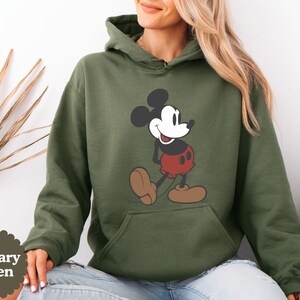 May include: Military green hoodie featuring a classic Mickey Mouse graphic. The cartoon character is depicted in his iconic red shorts, brown shoes, and black and white features. The hoodie has a kangaroo pocket and a drawstring hood.